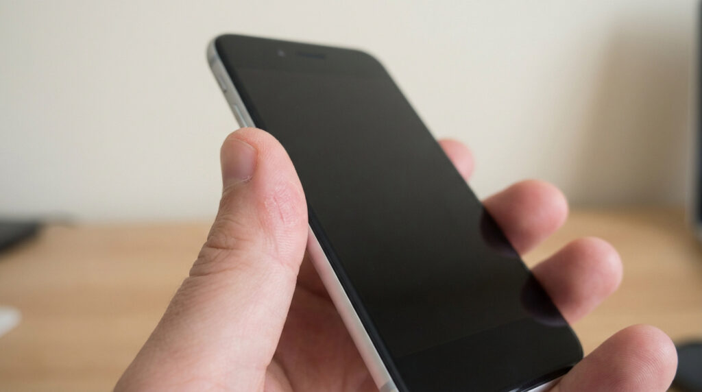 Main d'une personne tenant un smartphone noir à l'écran éteint. Le pouce présente des marques, suggérant un contact cutané régulier.