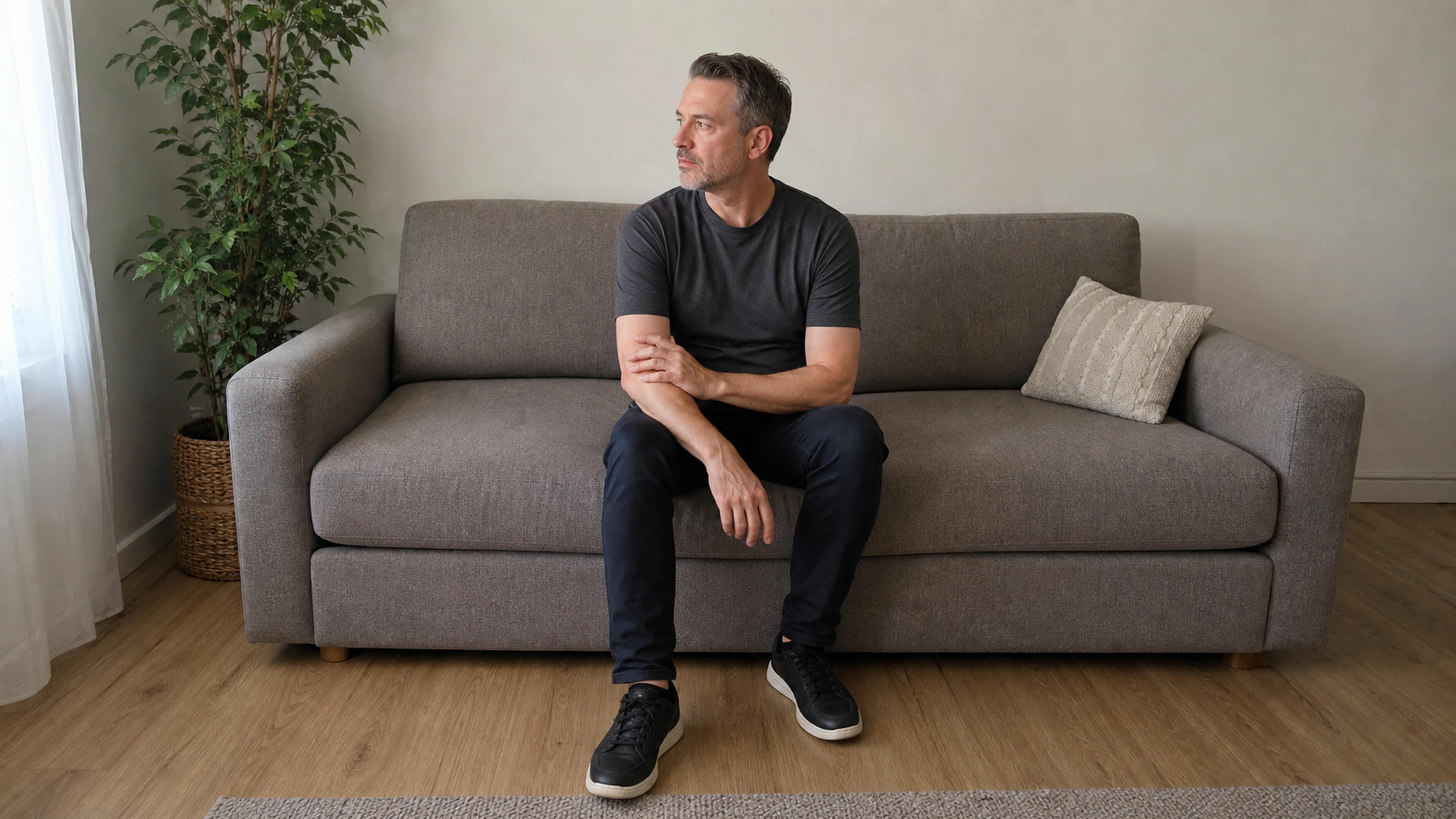 Homme assis touchant son avant-bras gauche, les deux jambes visibles dans un salon