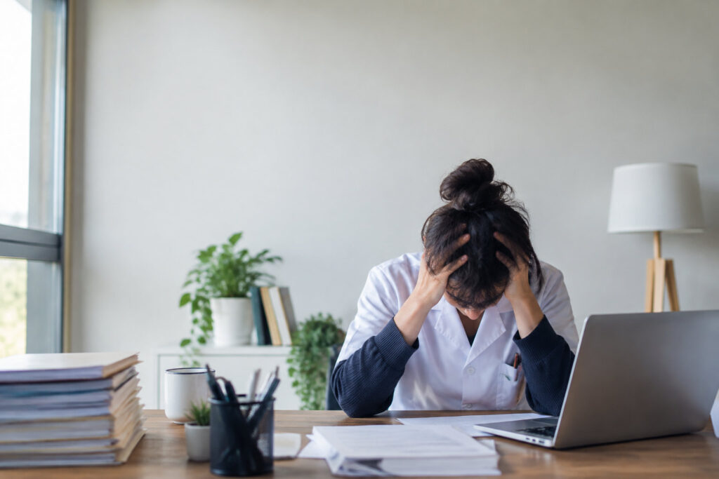 Une professionnelle de santé en blouse blanche, stressée, la tête dans les mains devant son ordinateur.
